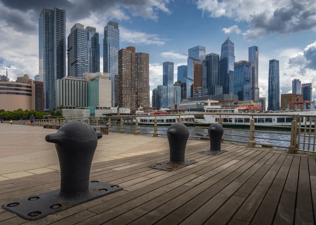 Wooden waterfront pier with cast-iron mooring bollards facing a modern city skyline and tour boats on the river