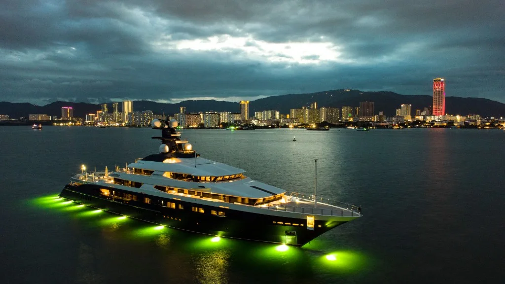 Luxury motor yacht with a dark hull and white decks at night, lit by warm cabin lights and green underwater lights, with a city skyline across the water
